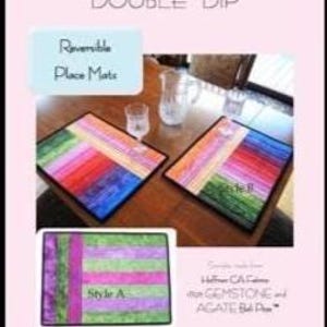 May include: Two reversible place mats with a colorful striped design, featuring shades of red, green, blue, and purple. The mats are displayed on a wooden table with a glass of water and a pitcher. The text "Auntie's Two Patterns Double Dip" is visible.
