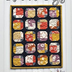 May include: A mini quilt titled "Lil' June Bugs" with a dark blue background and a yellow border. The quilt features sixteen colorful patchwork squares in various floral and geometric patterns. The Running Doe Quilts logo is at the bottom.
