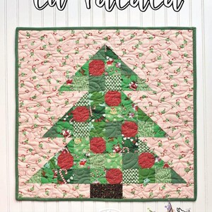 May include: A mini quilt with a Christmas tree design. The tree is made of green and red patterned fabric squares, with a pink background featuring a holly berry pattern. The quilt has a green border and the text "Lil' Falala" is at the top.