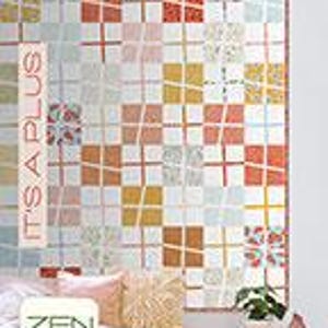 May include: A colorful quilt with a geometric pattern of squares in shades of orange, yellow, white, and blue. The quilt is displayed on a bed with pillows and a small side table. The text "IT'S A PLUS" is printed vertically on the left side of the quilt.