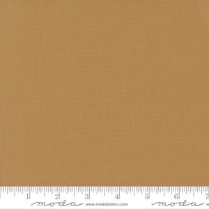 May include: Close-up of a solid, textured fabric in a warm, golden-brown hue. The fabric has a subtle woven pattern, and a ruler is placed along the bottom edge. The fabric is likely used for quilting or other sewing projects.
