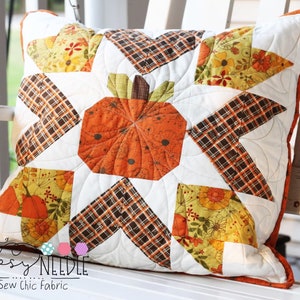 May include: Square decorative pillow with a quilted pumpkin design. The pillow features an orange pumpkin in the center, surrounded by white, plaid, and floral patterned fabric. The pillow has an orange border and is on a white surface.