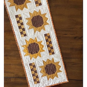 May include: A quilted table runner with a sunflower design. The runner features alternating sunflower and geometric block patterns in shades of brown, gold, and white. Finished size is 14.5" x 57.5". The text "Golden Glow" and "Table Runner Project" are visible.