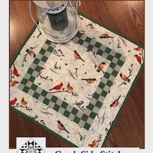 May include: A square quilted topper with a bird and branch pattern, featuring a green and white checkered border. The topper measures 20.5 x 20.5 inches. The text "Check It Out" and "Creek Side Stitches" are visible.