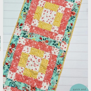May include: A rectangular quilted table runner featuring a patchwork design with coral, yellow, and teal floral patterns. The runner measures 16 inches by 72 inches and includes text reading The Tipsy Needle All Boxed Up Table Runner # TTN - 102.
