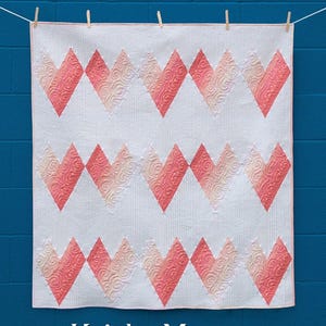 May include: A white baby quilt with a repeating pattern of pink and peach hearts. The quilt is suspended by clothespins against a blue brick wall. Text on the quilt reads "Mi Amor Baby Quilt And Table Runner" and "Krista Moser The Quilted Life". The finished size is 46.5" x 48.5".