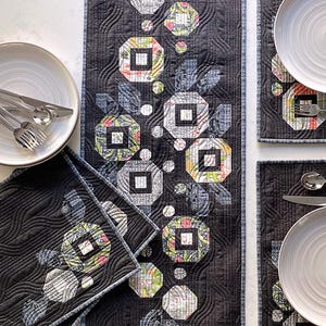 May include: A black quilted table runner and four placemats with a floral design. The table runner and placemats feature a repeating pattern of octagonal shapes with floral and geometric patterns. The text "POSIES" and "Table Runner and 4 Placemats" are visible.