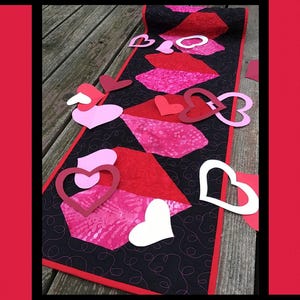 May include: A black and red quilted table runner with a heart design. The runner is surrounded by paper hearts in various colors. The text "Smitten" is at the top of the image. The text "Tricia Lynn Maloney Orphan Quilt Designs For Villa Rosa Designs" is at the bottom of the image.