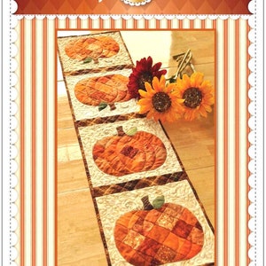 May include: A rectangular patchwork table runner featuring four square blocks, each displaying a fabric pumpkin in shades of orange, brown, and tan. The runner is bordered by a cream-colored fabric with a swirling quilted pattern. Text reads Shabby Fabrics and Patchwork Pumpkin Table Runner.