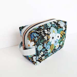May include: A floral print cosmetic bag with a black background and a variety of flowers in white, blue, and yellow. The bag has a striped handle and zipper, and a rose gold zipper pull.