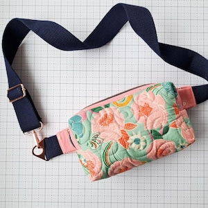 May include: A pink quilted fanny pack with a floral print and a navy blue adjustable strap. The strap has a silver clasp and a copper buckle.