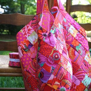 May include: A large, pink and red patchwork tote bag with floral and geometric patterns. The bag is made of fabric scraps sewn together in a diagonal pattern.