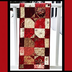 May include: A red, white, and brown patchwork quilt table runner with a "Ruby Road" title at the top. The quilt is hanging on a white railing. The text "Tricia Lynn Maloney Orphan Quilt Designs for Villa Rosa Designs" is at the bottom of the image.