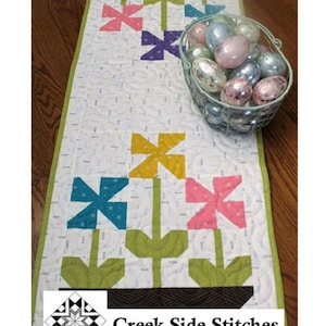 May include: A quilted table runner with a floral design. The runner is white with colorful pinwheel flowers in pink, yellow, blue, and purple. The runner is 15 inches by 40 inches. A wire basket filled with decorative eggs sits on the runner. The text "Spring Flowers" is at the top.