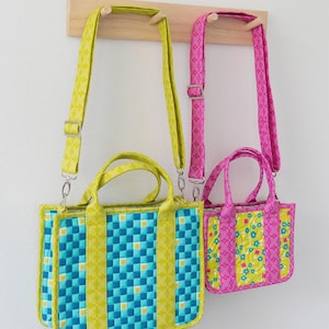 May include: Two crossbody tote bags hanging on a wooden rack. The larger bag has a blue and yellow checkered pattern, while the smaller bag has a floral design. The bags have matching straps and handles. Text at the top reads "Zuri Crossbody Tote".