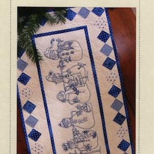 May include: A rectangular quilted table runner featuring blue embroidered snowman illustrations on a cream background. The border consists of alternating blue and white patterned fabric squares. The text "Snow Happens" is embroidered on a sign.