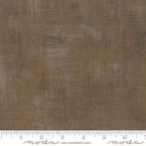 May include: A brown fabric with a textured, distressed look. The fabric has a subtle, mottled pattern with shades of brown and gray. The fabric is shown with a ruler for scale.