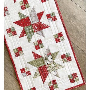 May include: A quilted table runner with a star pattern. The runner features red, green, and white fabrics. The design includes star shapes and small square blocks with the text "FA LA LA". Finished size is 14.5" x 44.5".