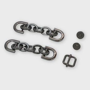 May include: A collection of dark gray metal hardware components, including two chain closures, two magnetic snaps, and a small buckle. The chain closures feature a series of interconnected rings and curved end pieces. The hardware is designed for use in bags or other accessories.