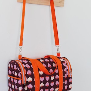 May include: A bright orange duffel bag with a strawberry print hanging on a wooden hook. The bag has a zipper closure and two orange straps. The text "Lucy Duffle" is at the top of the image and "Knot + Thread Design" is at the bottom.