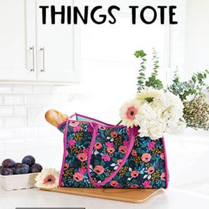May include: A floral tote bag with pink accents, filled with flowers and a baguette. The bag is on a wooden cutting board, with plums in a white dish. Text on the bag says "ALL THE THINGS TOTE".