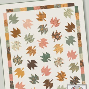 May include: A quilt with a cream background featuring numerous leaf shapes in various colors and patterns. The leaves are in shades of brown, green, pink, and yellow. The quilt is bordered with patterned fabric and has the text "Maple Valley" at the top.