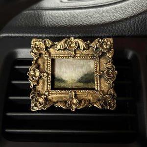 May include: A small, ornate gold-colored picture frame with a landscape painting inside. The frame is rectangular with detailed scrollwork and is placed on a black car vent.