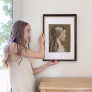 May include: A framed portrait of a young woman with long brown hair, wearing a white dress, against a green background. The portrait is hanging on a white wall.