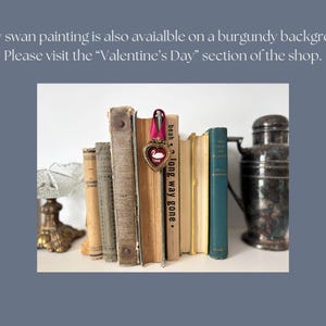 May include: A collection of vintage books with aged covers, some with visible titles. A heart-shaped ornament with a swan design hangs from a pink ribbon. A decorative metal container is on the right. The text in the image says: "A tiny swan painting is also available on a burgundy background."