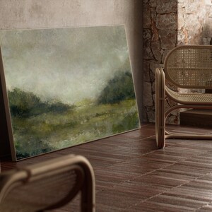A thin wooden frame holds a vintage landscape oil painting. The art is leaning against a stone wall beside modern cane chairs.