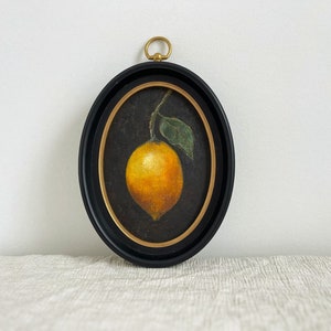 An oval black frame has a vintage still life art print of a lemon on a black background in it. The frame is on beige fabric leaning against a white wall.