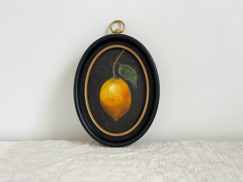 An oval black frame has a vintage still life art print of a lemon on a black background in it. The frame is on beige fabric leaning against a white wall.
