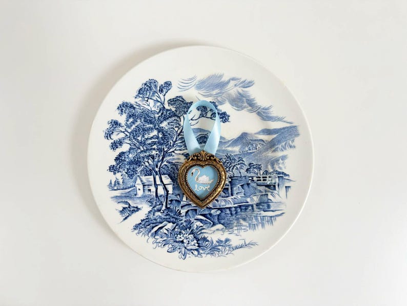 Tiny framed swan painting in a heart-shaped vintage gold frame with pale blue ribbon, displayed on blue and white toile china plate, romantic cottagecore decor.