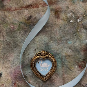 A miniature swan painting on patina blue in a vintage heart shaped tiny frame. An ice blue ribbon is lying on a paint stained surface.