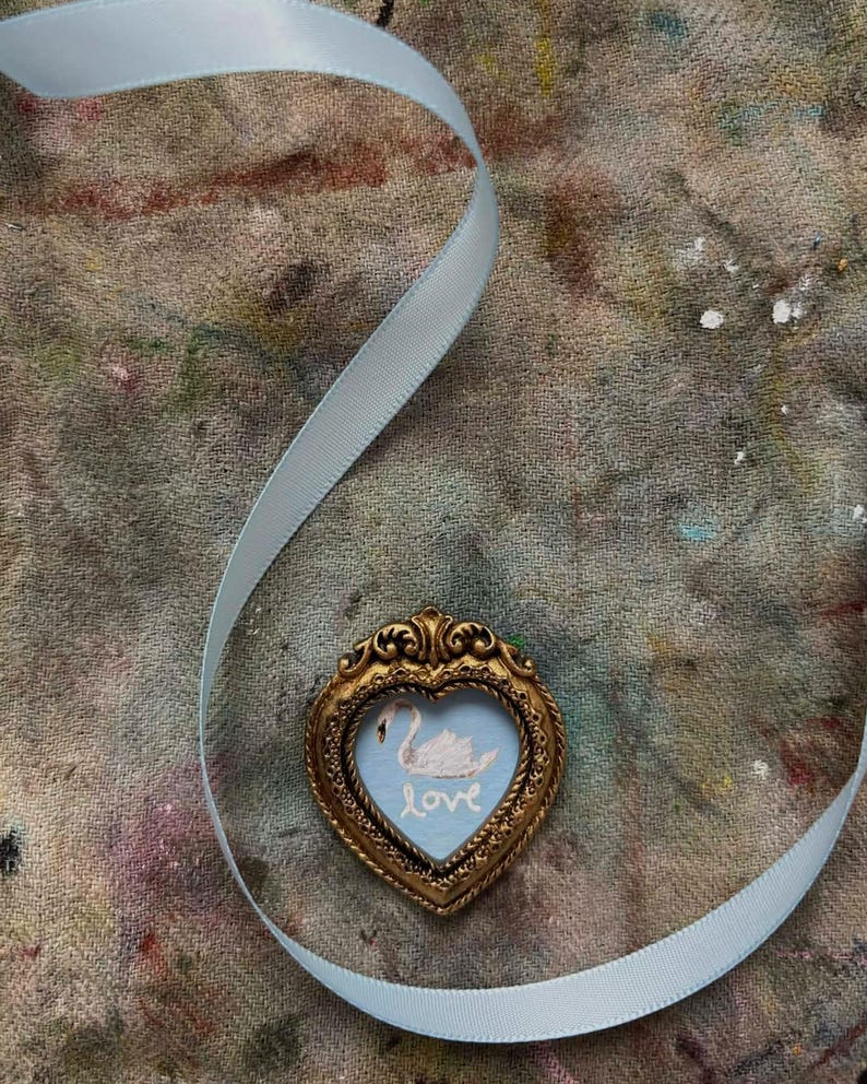 A miniature swan painting on patina blue in a vintage heart shaped tiny frame. An ice blue ribbon is lying on a paint stained surface.