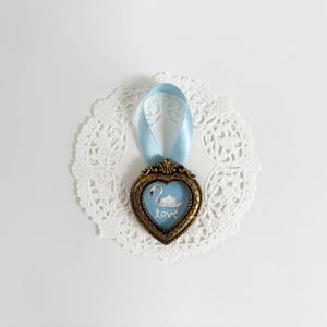 Tiny framed swan painting in a heart-shaped vintage gold frame with pale blue ribbon, displayed on on white lace doily, romantic cottagecore decor.