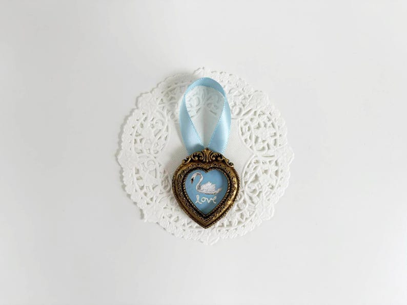 Tiny framed swan painting in a heart-shaped vintage gold frame with pale blue ribbon, displayed on on white lace doily, romantic cottagecore decor.