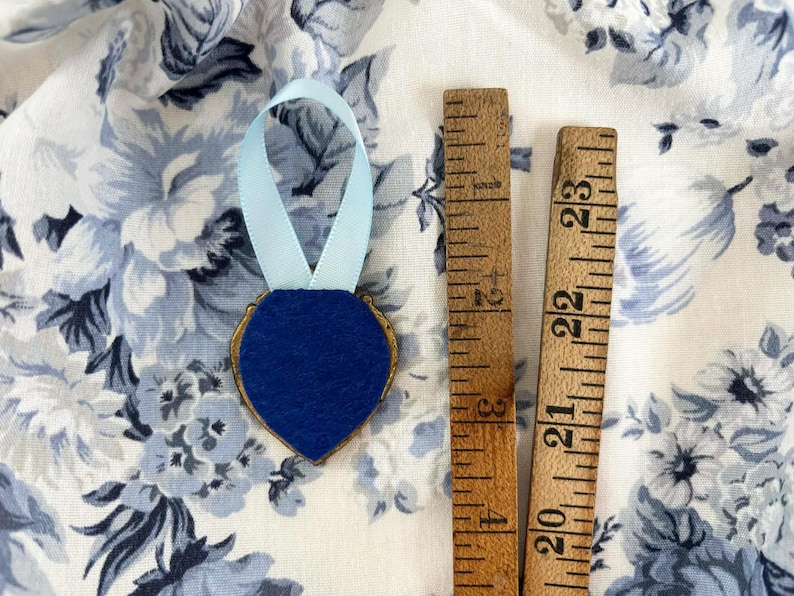 The back of a tiny framed painting in a heart-shaped vintage gold frame with pale blue ribbon and royal blue felt, displayed on blue and white roses, romantic cottagecore decor.