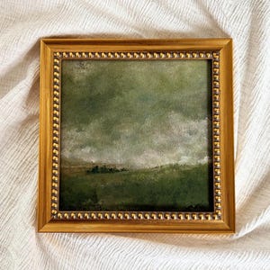 A gold frame with beading frames a vintage English countryside landscape art print. The frame is lying on beige fabric.