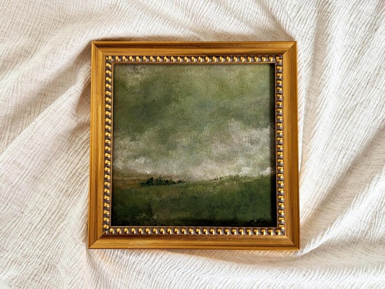 A gold frame with beading frames a vintage English countryside landscape art print. The frame is lying on beige fabric.