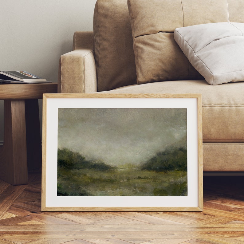 A large wooden frame with a white mat holds a vintage landscape oil painting. The painting is leaning against a modern brown couch.