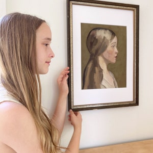 May include: A young person with long blonde hair is looking at a framed painting of a woman with long brown hair. The painting is in a brown frame with a gold trim. The woman in the painting is wearing a white shirt and has a neutral expression on her face.