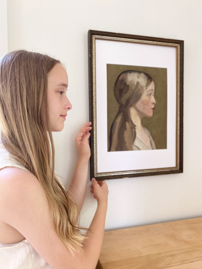 May include: A young person with long blonde hair is looking at a framed painting of a woman with long brown hair. The painting is in a brown frame with a gold trim. The woman in the painting is wearing a white shirt and has a neutral expression on her face.
