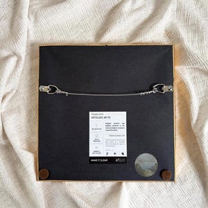 The back of a frame has a wire and hanging hooks. It lies on beige fabric.