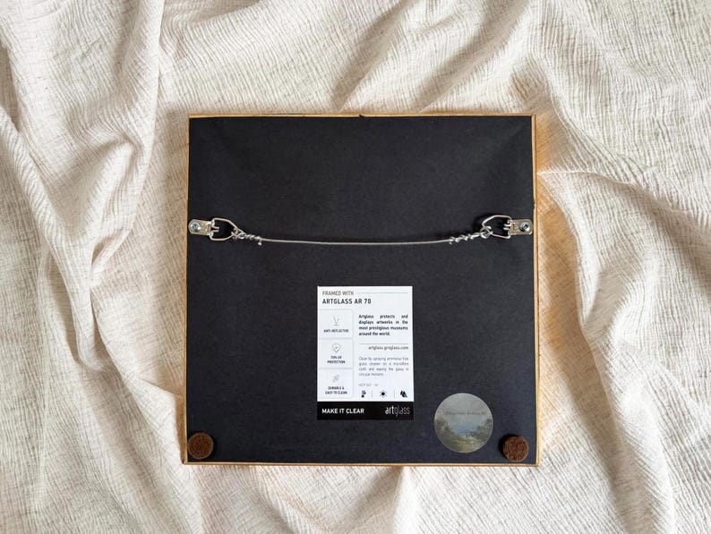 The back of a frame has a wire and hanging hooks. It lies on beige fabric.