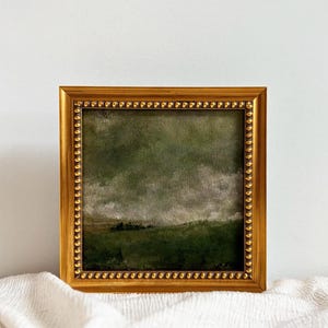 A gold frame with beading frames a vintage English countryside landscape art print. The frame is sitting on beige fabric with a white wall in the background.