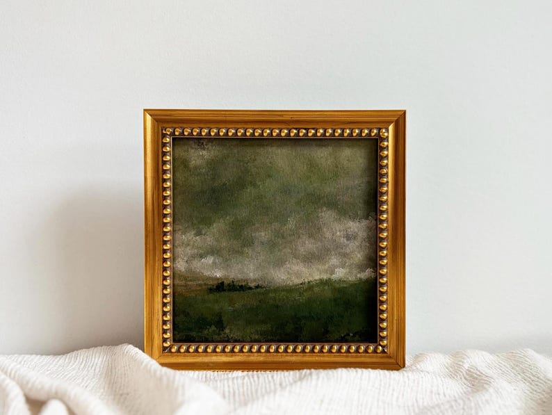 A gold frame with beading frames a vintage English countryside landscape art print. The frame is sitting on beige fabric with a white wall in the background.