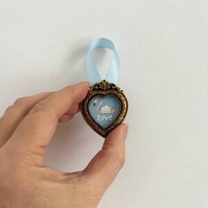 A caucasion hand is holding a tiny framed swan painting in a heart-shaped vintage gold frame with pale blue ribbon. The style is romantic cottagecore decor.