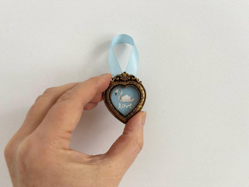 A caucasion hand is holding a tiny framed swan painting in a heart-shaped vintage gold frame with pale blue ribbon. The style is romantic cottagecore decor.