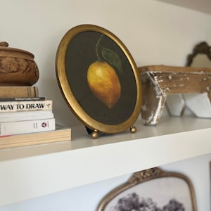 A lemon still life painting in a gold frame is on a shelf with antiques and vintage books.